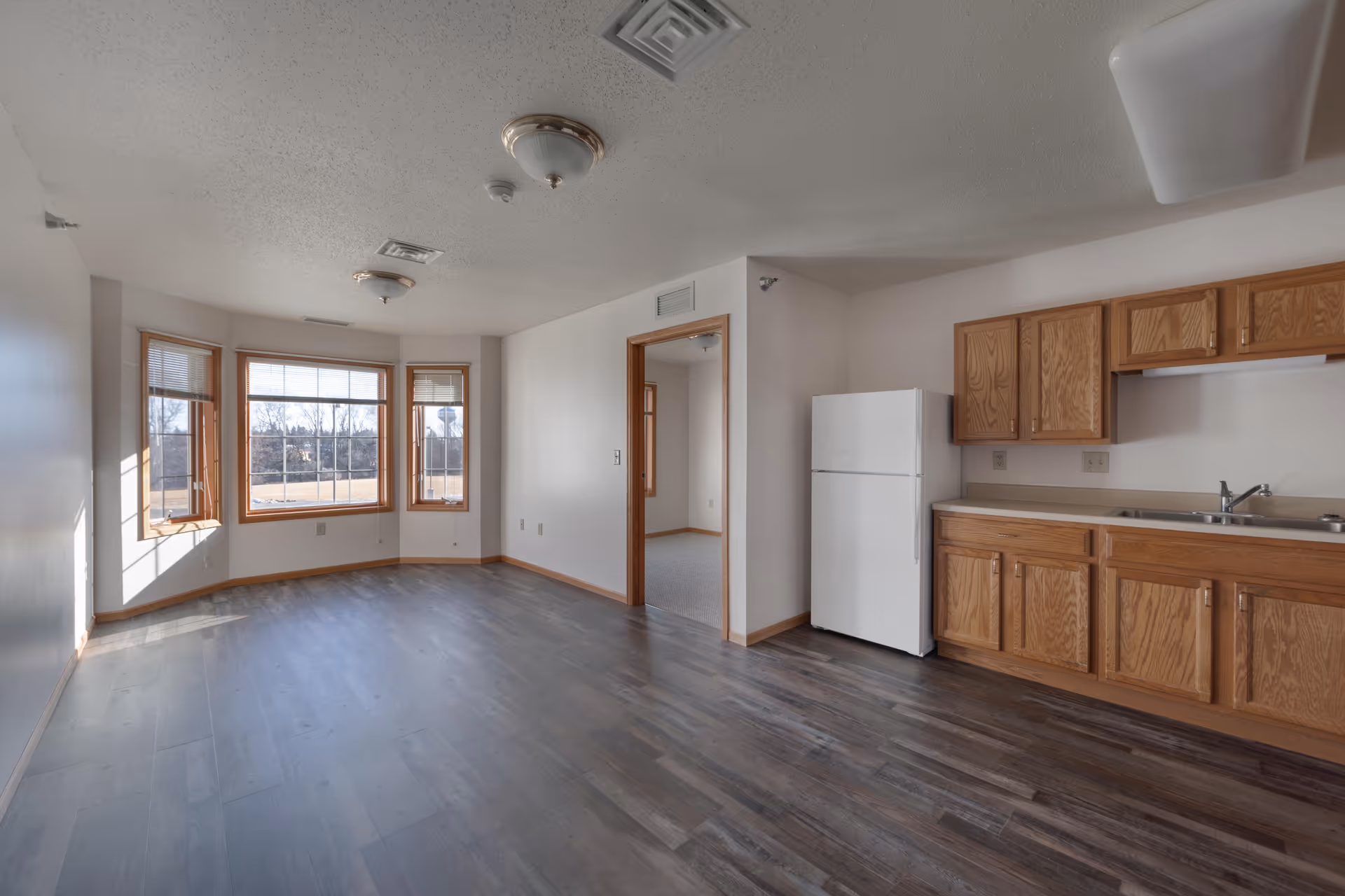 An empty room with a kitchen area featuring wooden cabinets, a white refrigerator, and a sink. The room has large windows with wooden frames letting in natural light, and dark wood flooring. There is an open doorway leading to another room with carpeted flooring.