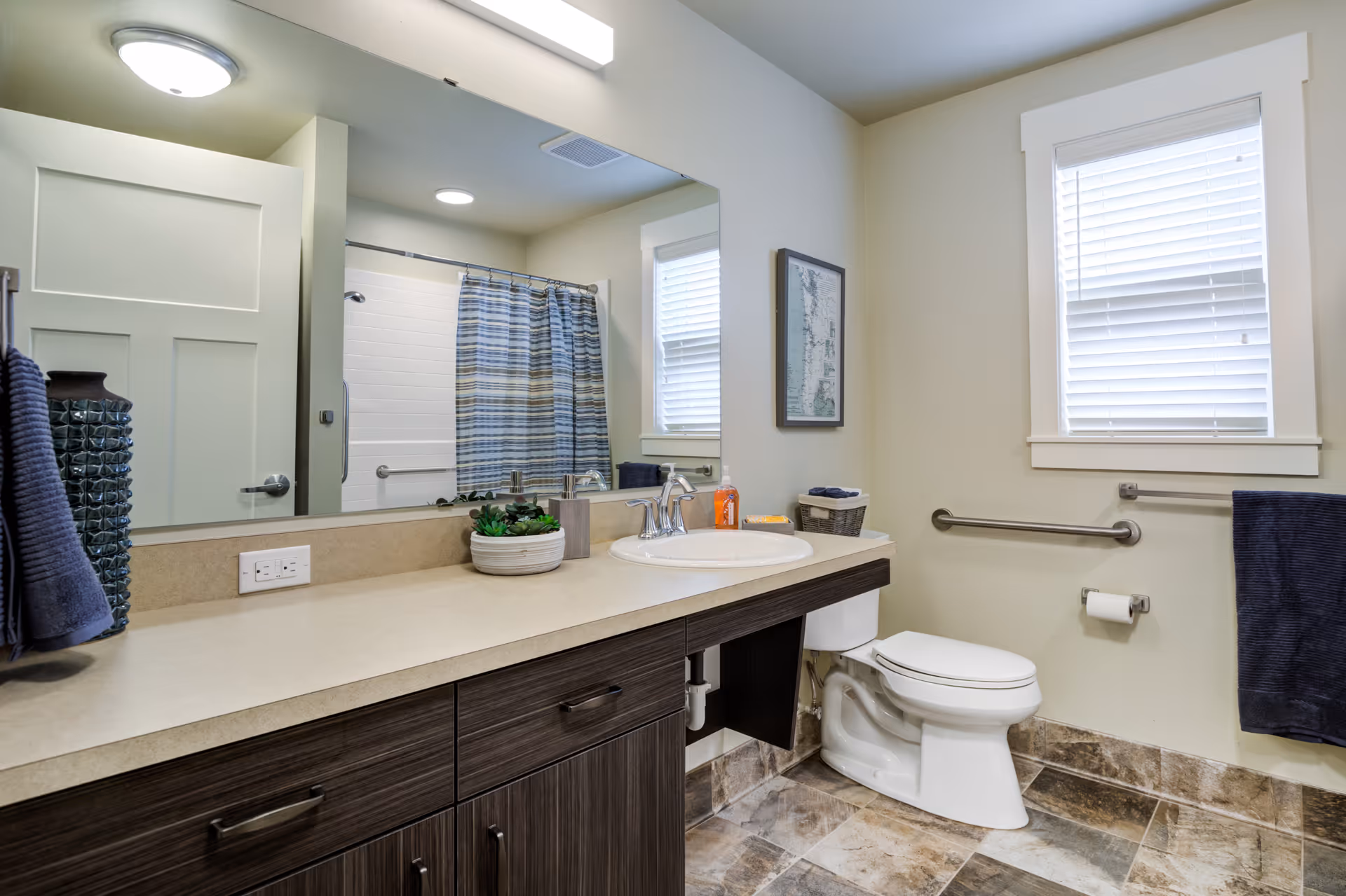 A clean and modern bathroom with a large mirror above a countertop with a sink. The bathroom features a toilet, a window with blinds, a towel rack with a dark blue towel, and a shower with a striped shower curtain. The floor is tiled, and there is a small plant and soap dispenser on the countertop.