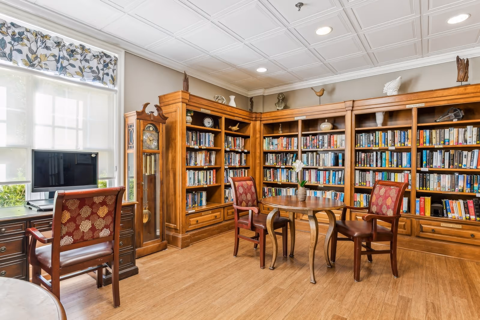 A cozy library room with wooden bookshelves filled with books, a round wooden table with a small vase of flowers, and three wooden chairs with red patterned upholstery. There is a grandfather clock near the window, which has floral-patterned curtains and lets in natural light. A computer monitor is placed on a wooden desk by the window.