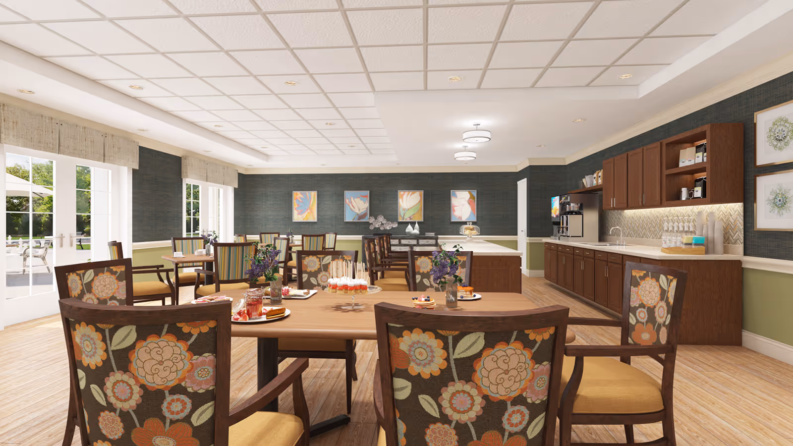 A bright and spacious dining room with wooden tables and chairs featuring floral upholstery. The room has large windows and glass doors letting in natural light, a long counter with cabinets and a coffee station along one wall, and colorful floral artwork on the walls. There are plates with desserts and flowers on the tables.