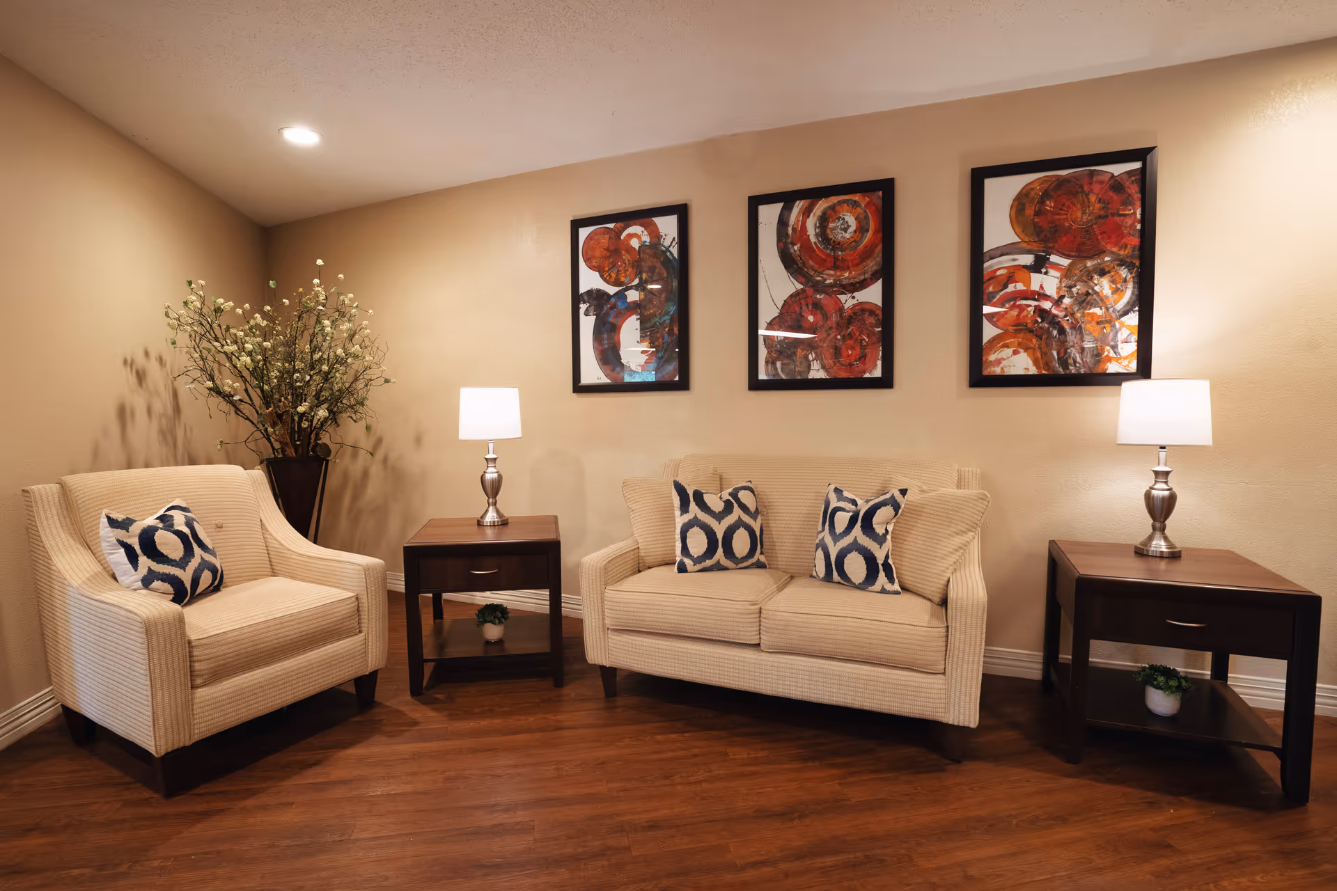 A cozy living room area with a beige armchair and a matching two-seater sofa, each adorned with patterned throw pillows. There are two dark wooden side tables with silver lamps on top, and a small potted plant on the lower shelf of each table. Behind the sofa, three abstract art paintings with red and orange tones hang on a beige wall. A tall vase with white flowers is placed in the corner. The floor is wooden.