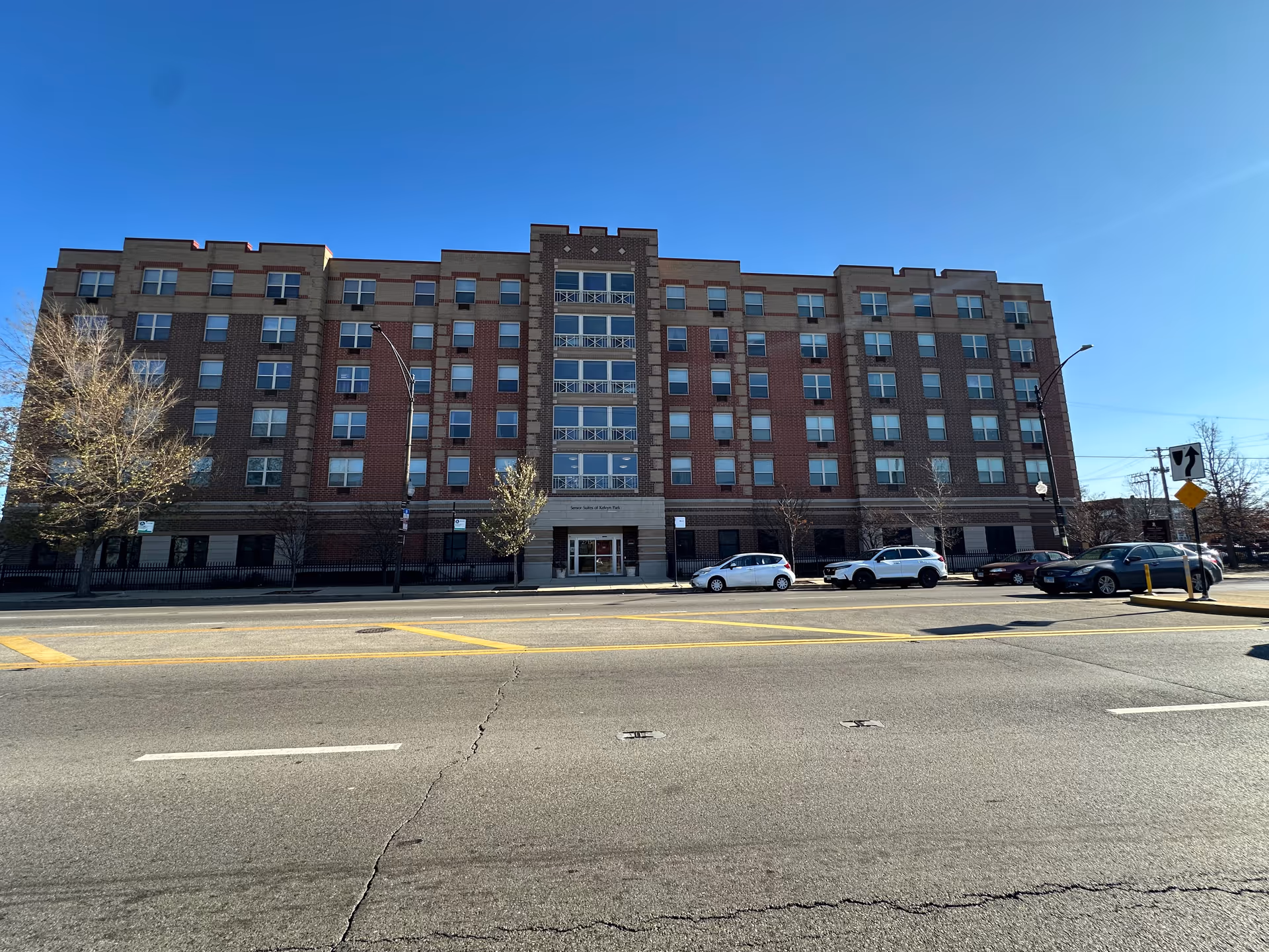 Exterior front view of a multi-story brick building named Senior Suites of Kelvyn Park with several windows, a central entrance, parked cars along the street, and a clear blue sky.