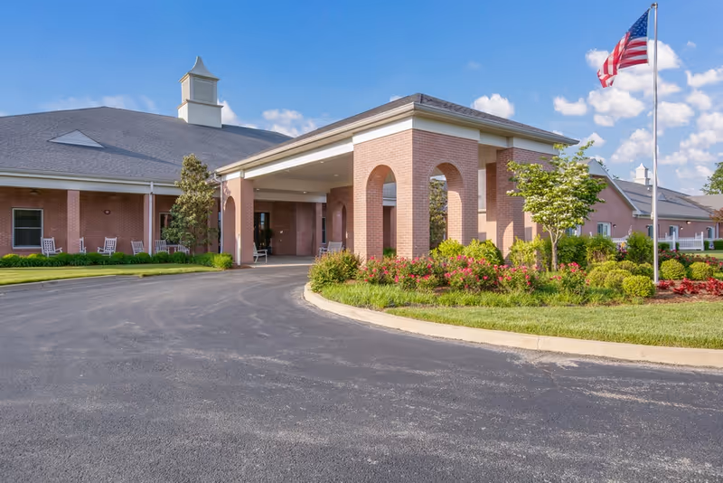 Exterior view of Inspirations of Mount Washington facility showing a brick building with a covered entrance, landscaped garden with flowers and shrubs, an American flag on a flagpole, and a clear blue sky with some clouds.