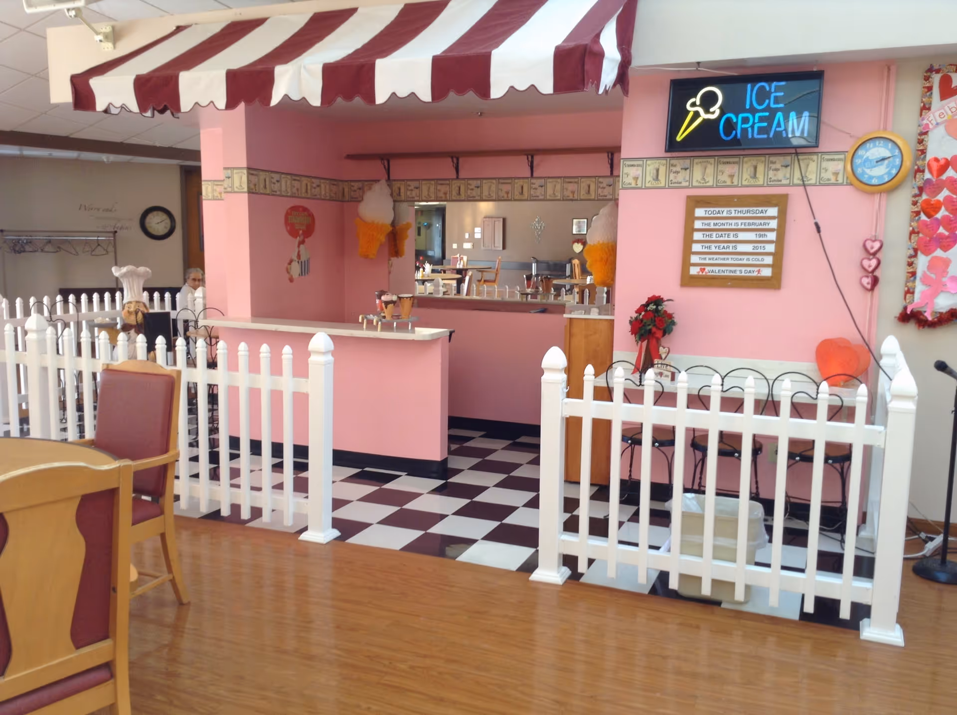 An indoor ice cream parlor area within a senior living facility featuring pink walls, a red and white striped awning, and a white picket fence surrounding the counter. The floor inside the fenced area has a black and white checkered pattern. There is a neon sign that reads 'ICE CREAM' with an ice cream cone graphic above the counter. A bulletin board displays the date and weather information, and there are heart decorations on the wall. Wooden chairs and tables are visible in the foreground.