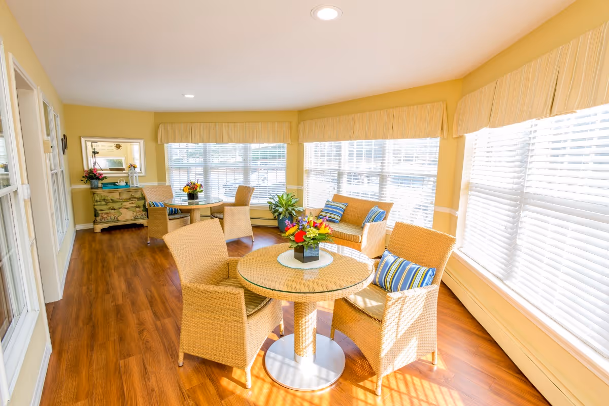 A bright and cozy sitting area with large windows covered by white blinds and yellow valances. The room features wooden flooring, round wicker tables with matching wicker chairs, and a wicker loveseat with striped cushions. There are colorful flower arrangements on the tables and a decorative chest with a mirror and plants against the wall.