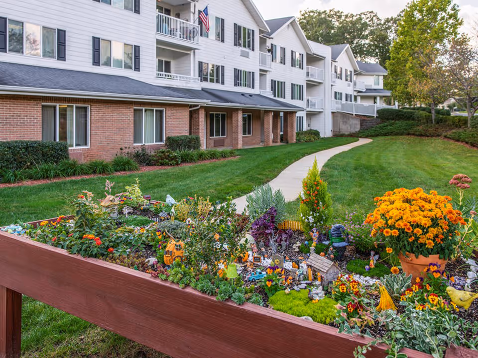 Raised flower bed filled with colorful flowers and miniature decorations in front of a multi-story residential building with balconies and a curved sidewalk.
