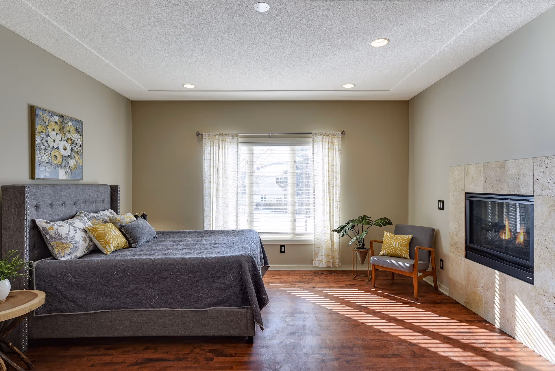 Bright, furnished bedroom featuring a bed with decorative pillows, a chair by a fireplace, and a window with sheer curtains.