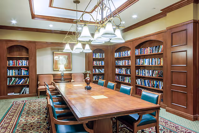 A well-lit library or meeting room with a long wooden table surrounded by green cushioned chairs. The walls are lined with wooden bookshelves filled with books. There are two upholstered armchairs and a table lamp in the corner, and a decorative chandelier hangs from the ceiling.