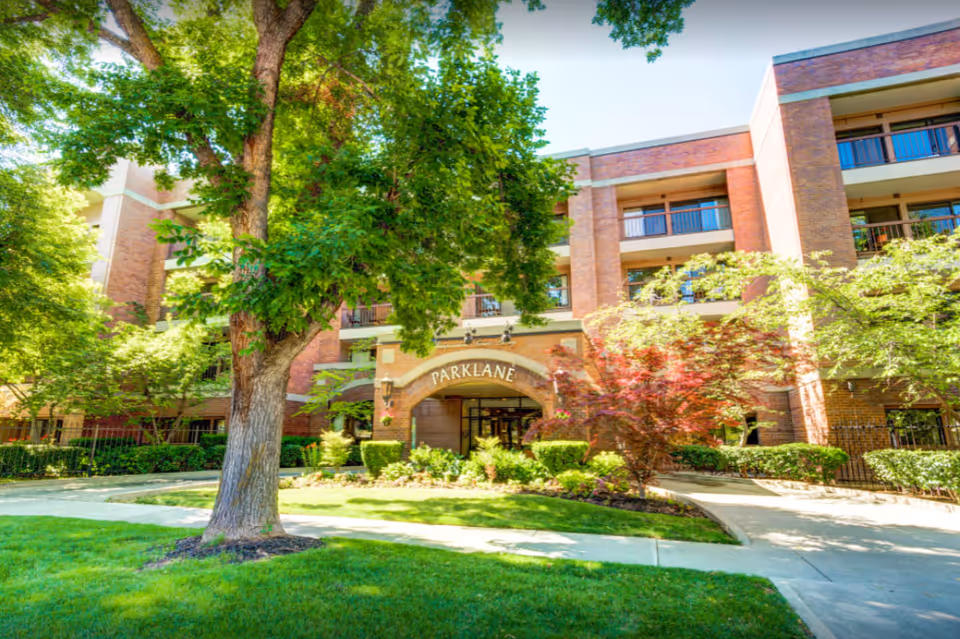 Exterior view of Park Lane Senior Living building with a large tree and well-maintained landscaping in front, including green grass, bushes, and a red-leafed tree. The building is made of brick and has balconies on the upper floors.