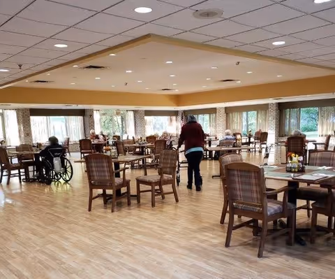 A spacious dining room in a senior living facility with wooden tables and chairs arranged neatly. Several elderly residents are seated at the tables, and one person is walking through the room. Large windows with curtains allow natural light to fill the space.