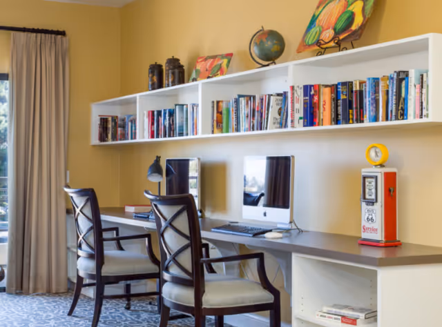 A bright communal computer workspace with two iMacs, chairs, wall-mounted bookshelves and decorative items.