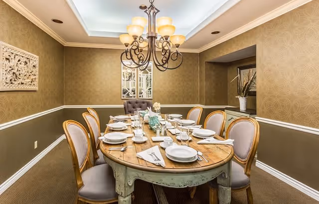 A formal dining room with a wooden oval table set for eight people with white plates, napkins, and glassware. The room features beige patterned wallpaper, a decorative wall panel, a chandelier with multiple lights, and a mirror on the far wall. There are upholstered chairs around the table and a small alcove with a vase of tall plants.