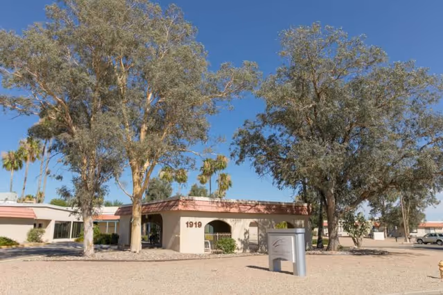 Exterior view of Casas Adobes Post Acute Rehabilitation Center building with beige walls and a red-tiled roof, surrounded by large trees and a gravel landscape under a clear blue sky. The building number 1919 is visible on the wall, and there is a sign in front of the building.