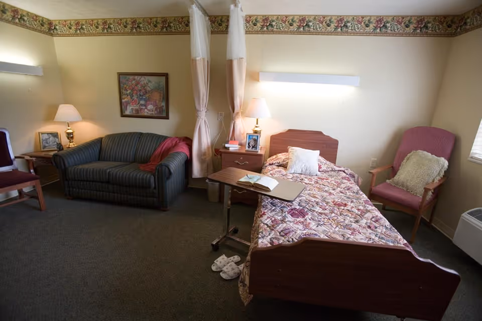 A cozy room with a single bed covered in a floral quilt, a small overbed table with an open book, a pair of slippers on the floor, a striped couch with a red blanket, two armchairs with cushions, two bedside tables with lamps, and a framed floral painting on the wall. The room has beige walls with a floral border near the ceiling and carpeted floor.