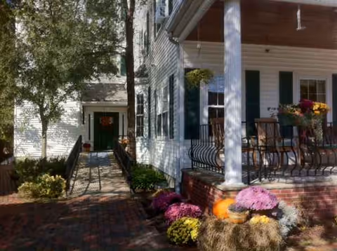 White two-story building with a covered porch, potted flowers, and a brick walkway