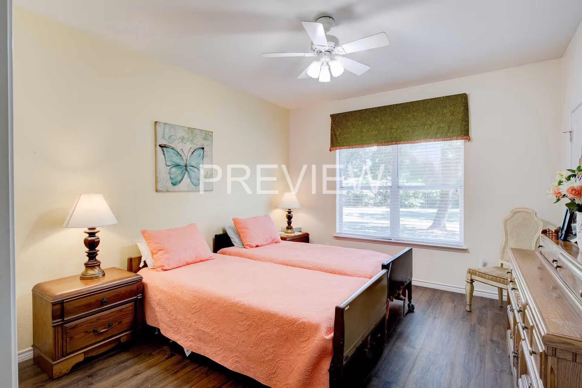 A bedroom with two single beds placed side by side, each covered with a pink bedspread and matching pillows. There are two wooden nightstands with lamps on either side of the beds. A large window with a green valance allows natural light to fill the room. A wooden dresser with a chair and a vase of flowers is positioned against the right wall. A ceiling fan with lights is mounted on the ceiling, and a butterfly artwork hangs on the wall above the beds.