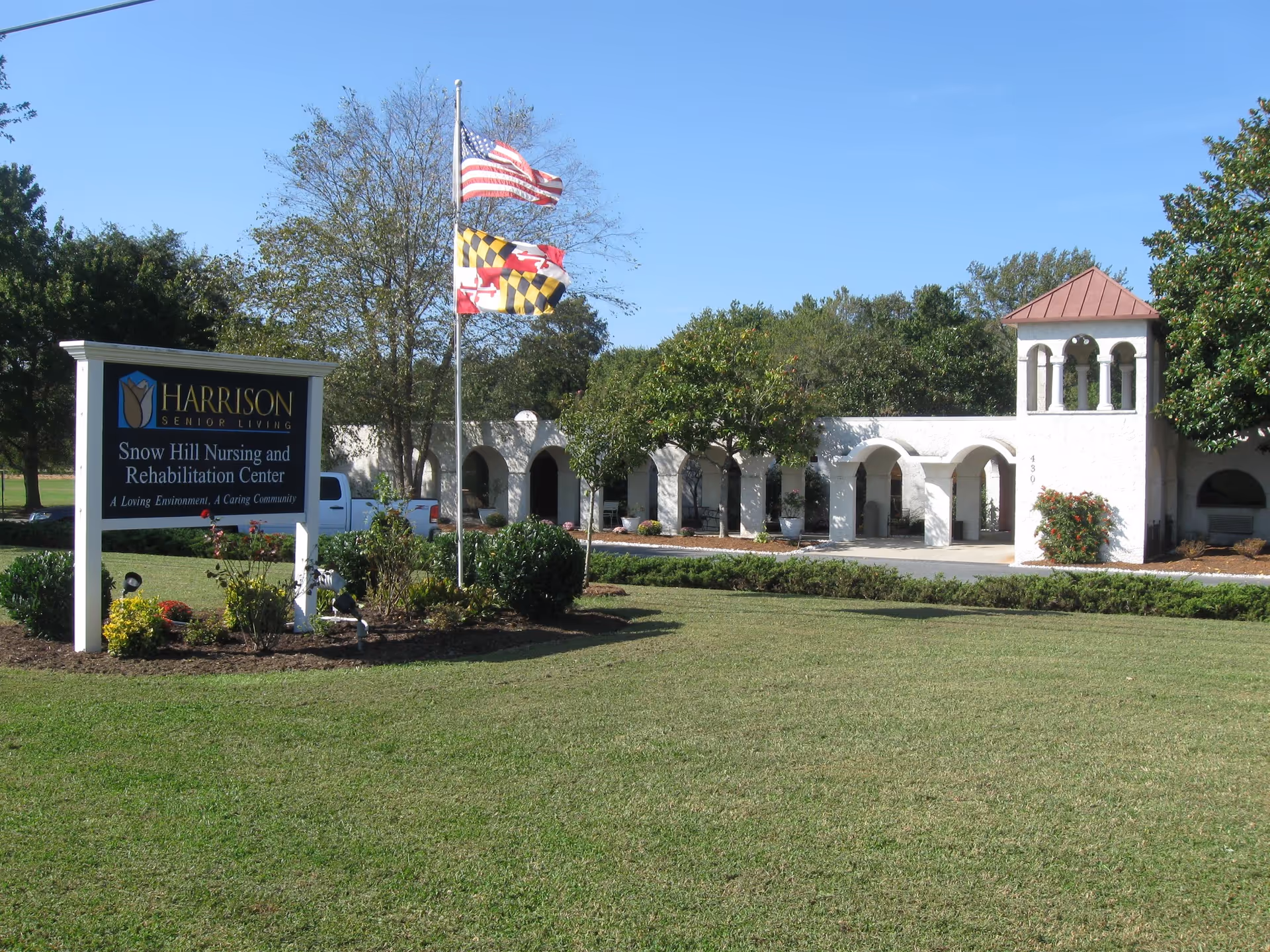 Exterior view of Snow Hill Nursing and Rehabilitation Center with a white building featuring arches and a small tower. There is a sign in the foreground that reads 'Harrison Senior Living Snow Hill Nursing and Rehabilitation Center A Loving Environment, A Caring Community'. Two flags, the American flag and the Maryland state flag, are flying on a flagpole near the sign. The area is landscaped with grass, bushes, and trees under a clear blue sky.