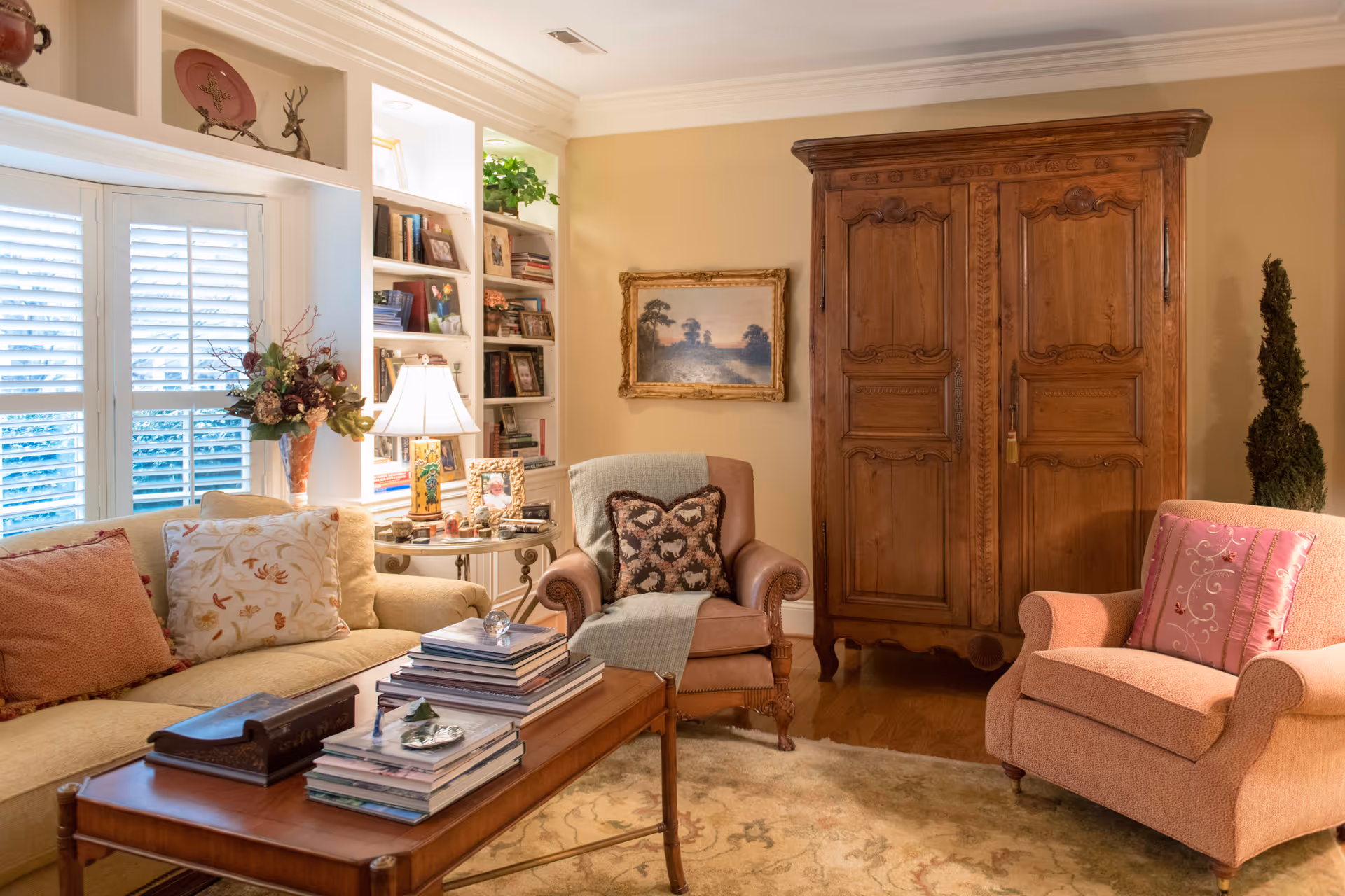 A cozy living room with a beige sofa adorned with patterned pillows, a wooden coffee table stacked with books, two upholstered armchairs with decorative cushions, a wooden armoire, built-in bookshelves filled with books and framed photos, a table lamp, and a window with white plantation shutters letting in natural light.