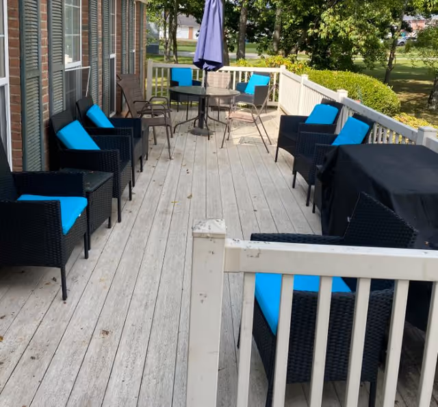 Outdoor patio area with multiple black wicker chairs featuring bright blue cushions arranged along a wooden deck. A round table with a closed purple umbrella and additional metal chairs are visible at the far end. The patio is surrounded by a white railing and overlooks a grassy area with trees.
