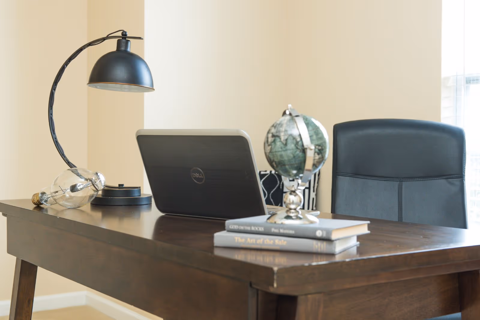 A wooden desk with a black desk lamp, a closed Dell laptop, a small globe on a stand, and two stacked books titled 'God on the Rocks' and 'The Art of the Sale'. Behind the desk is a black office chair and a window with blinds.