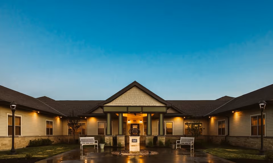 Front entrance of a single-story memory care building at dusk with a lit porch, benches, and lamp posts.