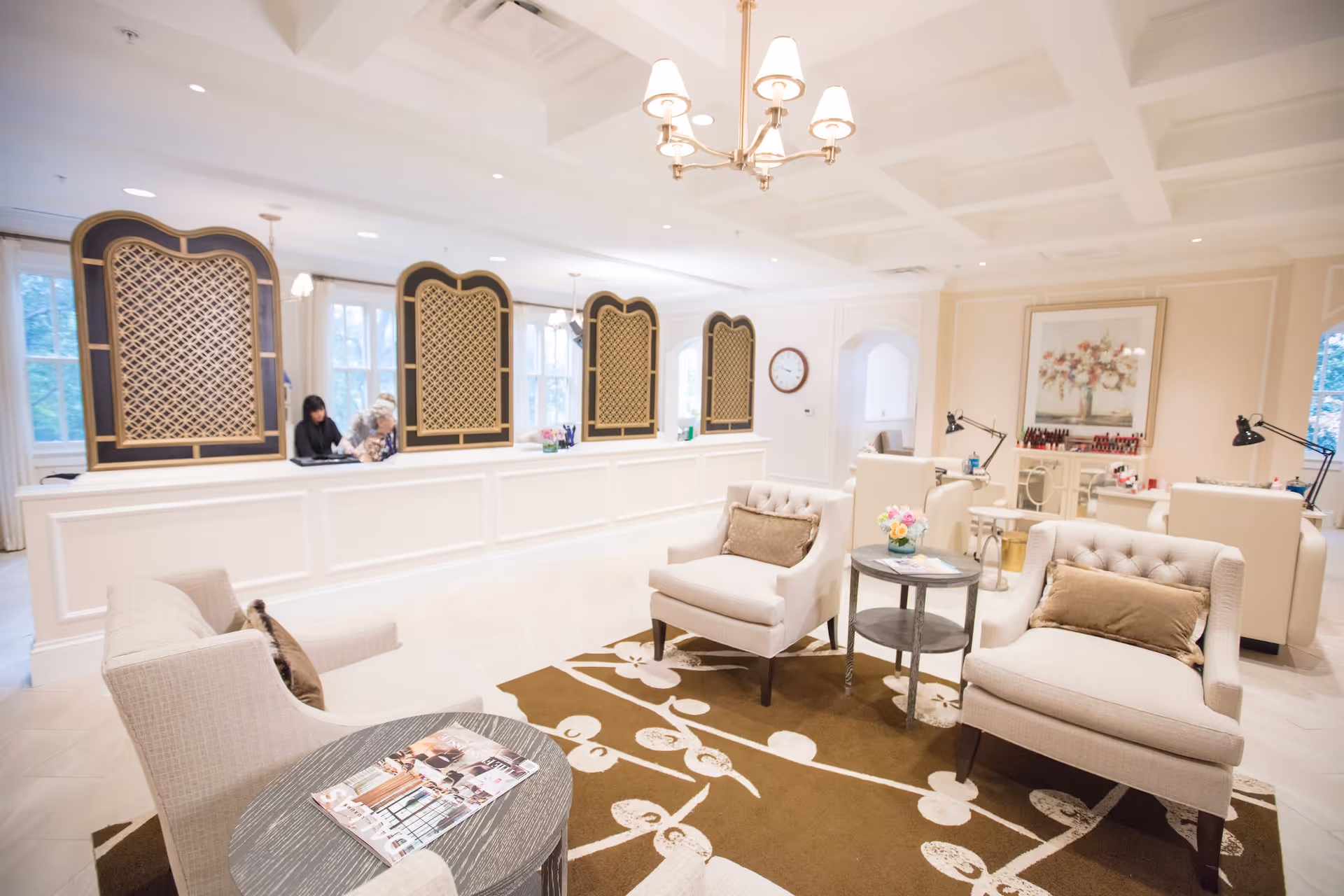 A bright and elegant reception area with a white counter featuring decorative lattice panels. Two women are seated behind the counter. In the foreground, there are comfortable beige armchairs with brown cushions arranged around a small round table on a brown and white patterned rug. The room has soft lighting with a chandelier and large windows letting in natural light. A wall clock and a floral painting are visible in the background.