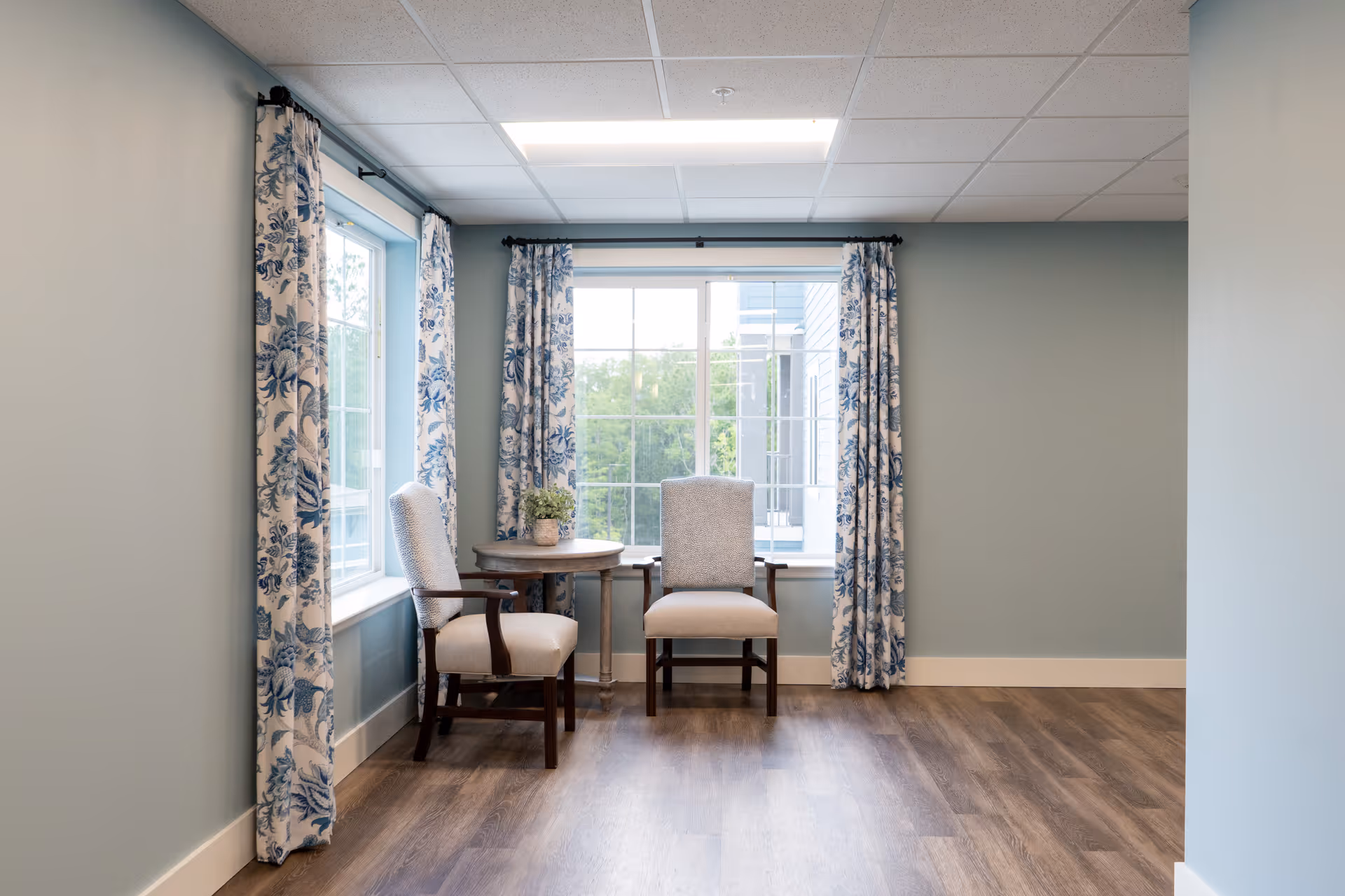 A small seating area with two upholstered chairs and a round wooden table between them, placed in front of two large windows with blue and white patterned curtains. The room has light blue walls and wood flooring.