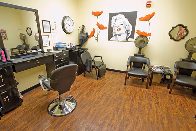 Interior view of a salon area with a styling chair in front of a mirror and counter with hair products. There are three black chairs along the wall, a small table with magazines, and wall decorations including a large black and white portrait of Marilyn Monroe with red lips and red poppy flower decals on the wall.