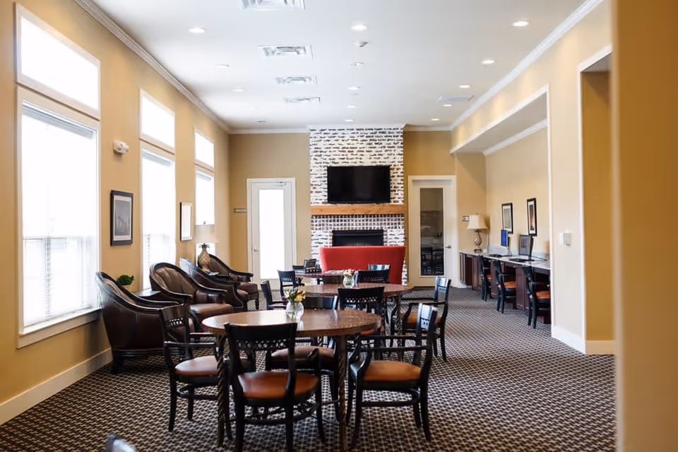 A well-lit common area with multiple round tables and chairs arranged on a patterned carpet. There are several leather armchairs along the left wall with large windows letting in natural light. At the far end, there is a white brick fireplace with a mounted flat-screen TV above it and a red couch in front. On the right side, there is a long desk with computers and chairs. The walls are painted beige with framed artwork and lamps.
