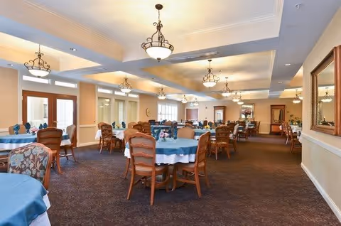 A spacious dining room with multiple round tables covered with blue tablecloths and wooden chairs. The room is well-lit with elegant ceiling lights and has large windows allowing natural light. The walls are painted in a light beige color, and there is a large mirror on the right wall.