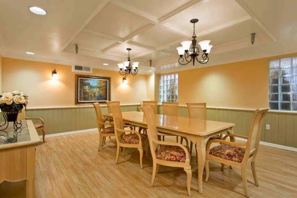 A dining room with a long wooden table surrounded by eight wooden chairs with floral upholstered seats. The room has light yellow walls with green wainscoting, two chandeliers hanging from a coffered ceiling, a framed painting on the wall, and glass block windows letting in natural light. A sideboard with a vase of flowers is visible on the left.