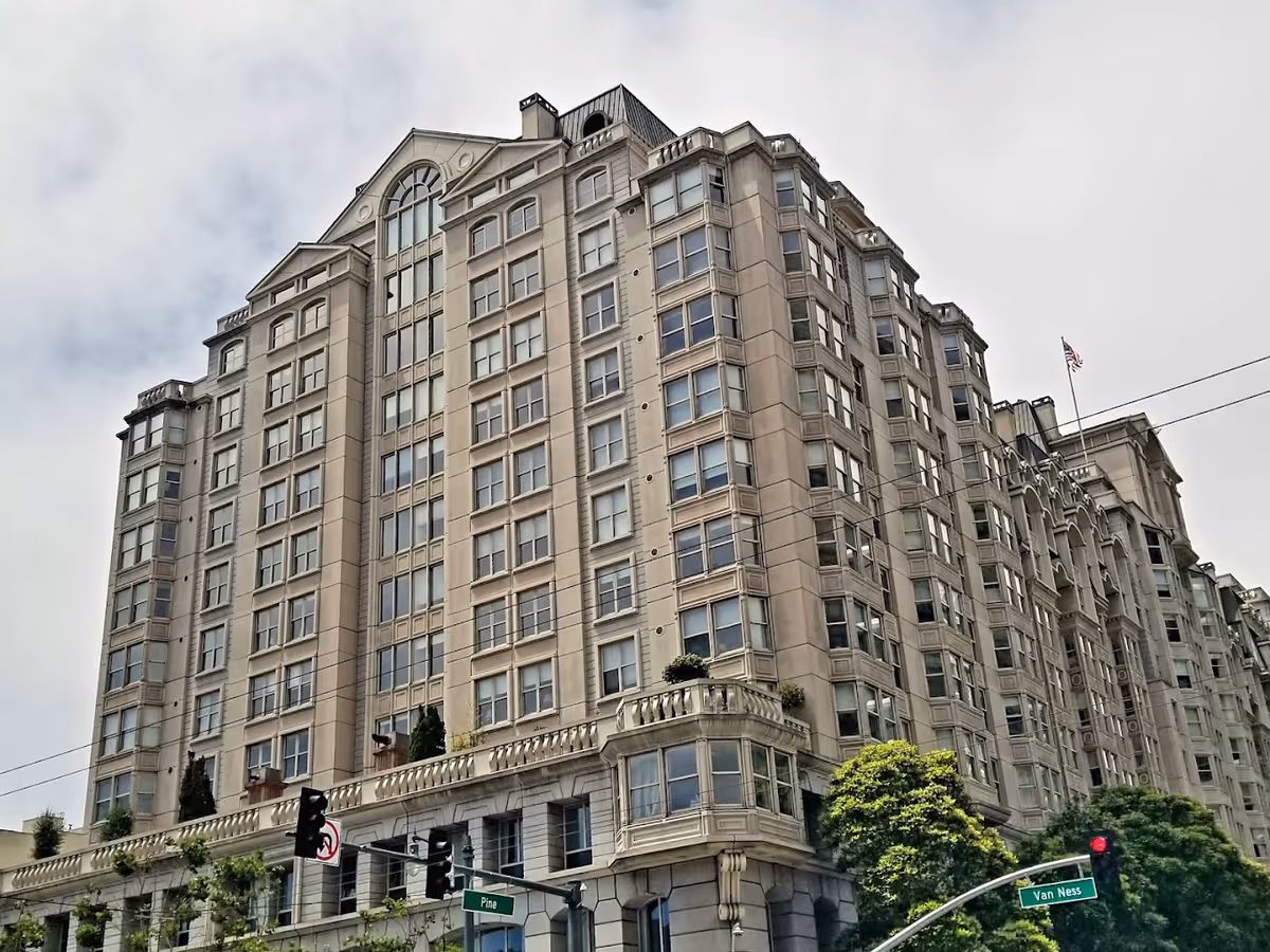 Exterior view of a large multi-story residential building with classical architectural details, numerous windows, and some greenery at the base. Traffic lights and street signs for Pine and Van Ness streets are visible in the foreground under a cloudy sky.