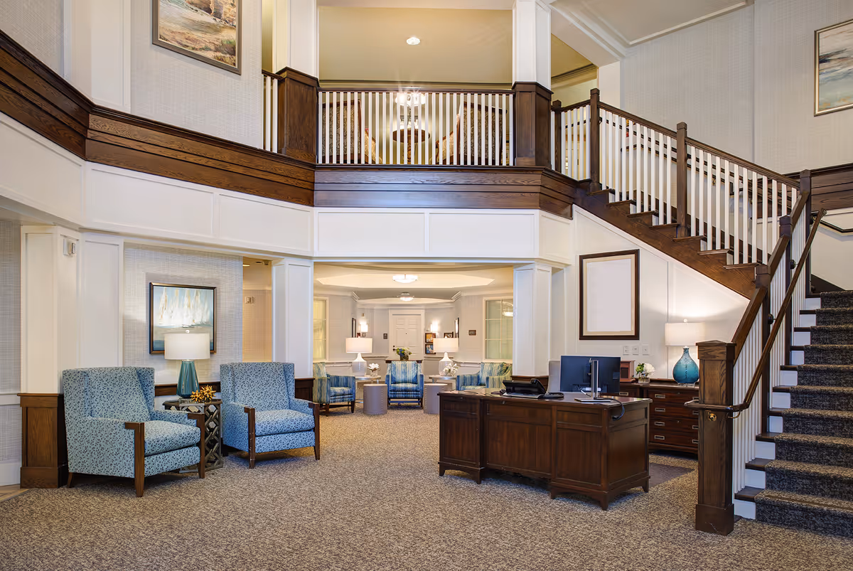 Lobby of a senior living facility with blue upholstered chairs, a wooden reception desk, and a staircase to an upper level.