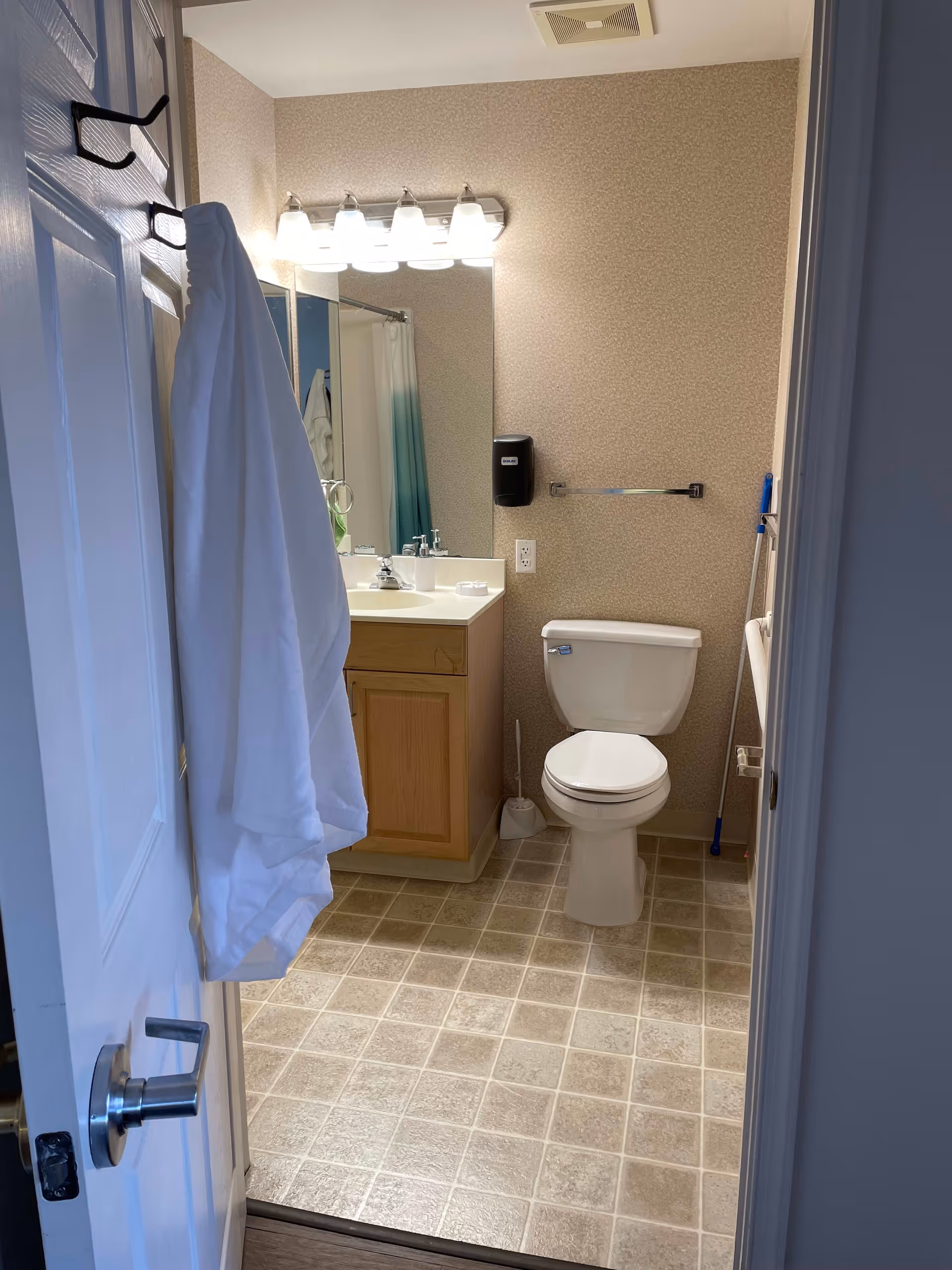 View into a bathroom with a white door partially open. A white bathrobe hangs on the door. Inside, there is a toilet, a wooden vanity with a sink, a large mirror above the sink with four lights, a soap dispenser mounted on the wall, and a toilet brush on the floor. The floor is covered with beige and light brown tiles.
