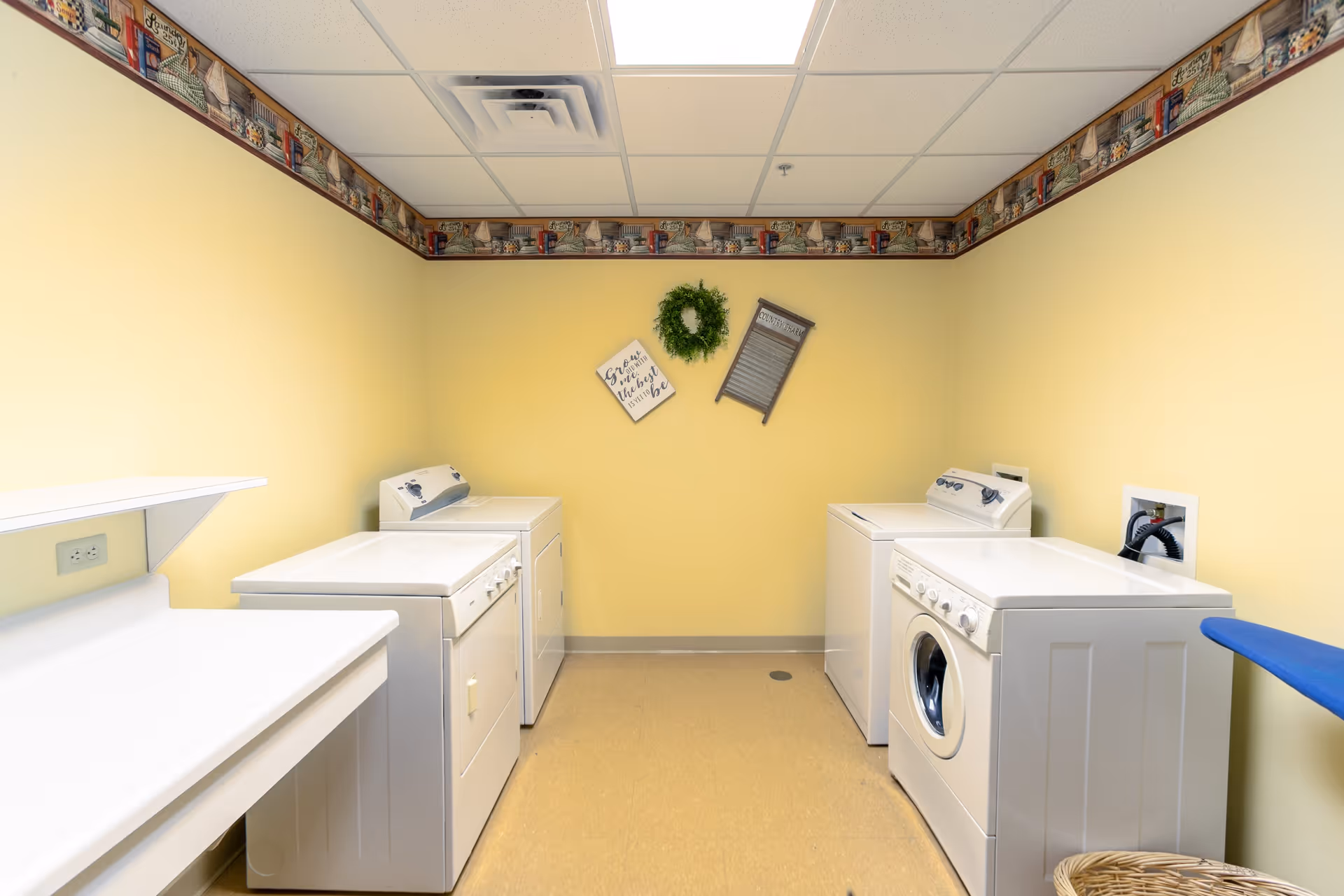 A laundry room with two washing machines and two dryers against yellow walls. There is a decorative border near the ceiling, a green wreath, a small sign, and a washboard hanging on the wall. An ironing board and a wicker basket are visible on the right side.