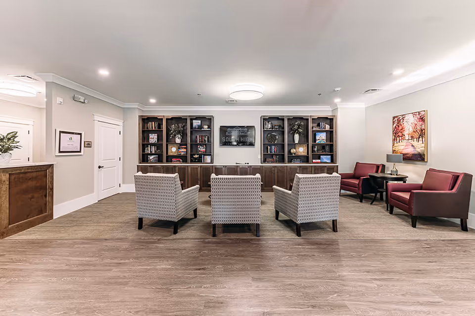 Bright communal lounge with three patterned armchairs facing built-in shelving and a central TV, with additional seating and artwork to the side.