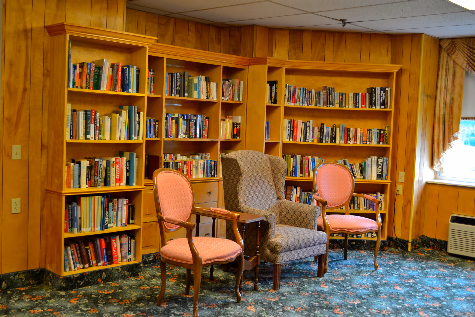 A cozy reading area in a senior living facility featuring wooden bookshelves filled with books, three chairs including a cushioned armchair and two wooden chairs with pink upholstery, a small wooden side table, floral patterned carpet, wood-paneled walls, and a window with curtains letting in natural light.
