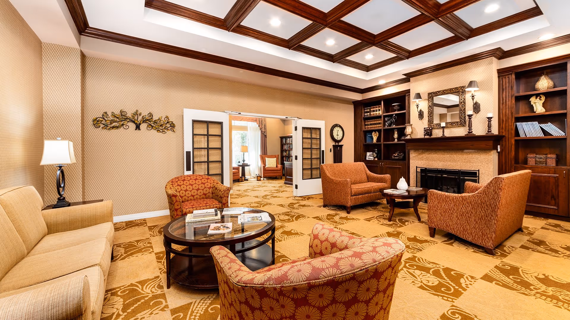 A spacious living room with beige and patterned carpet, featuring a beige sofa, two patterned armchairs, and two orange armchairs arranged around two wooden coffee tables. The room has a coffered ceiling with recessed lighting, a fireplace with a mirror and decorative items above it, and built-in wooden bookshelves on either side. A wall clock and a decorative wall piece are also visible, with double doors leading to another room in the background.