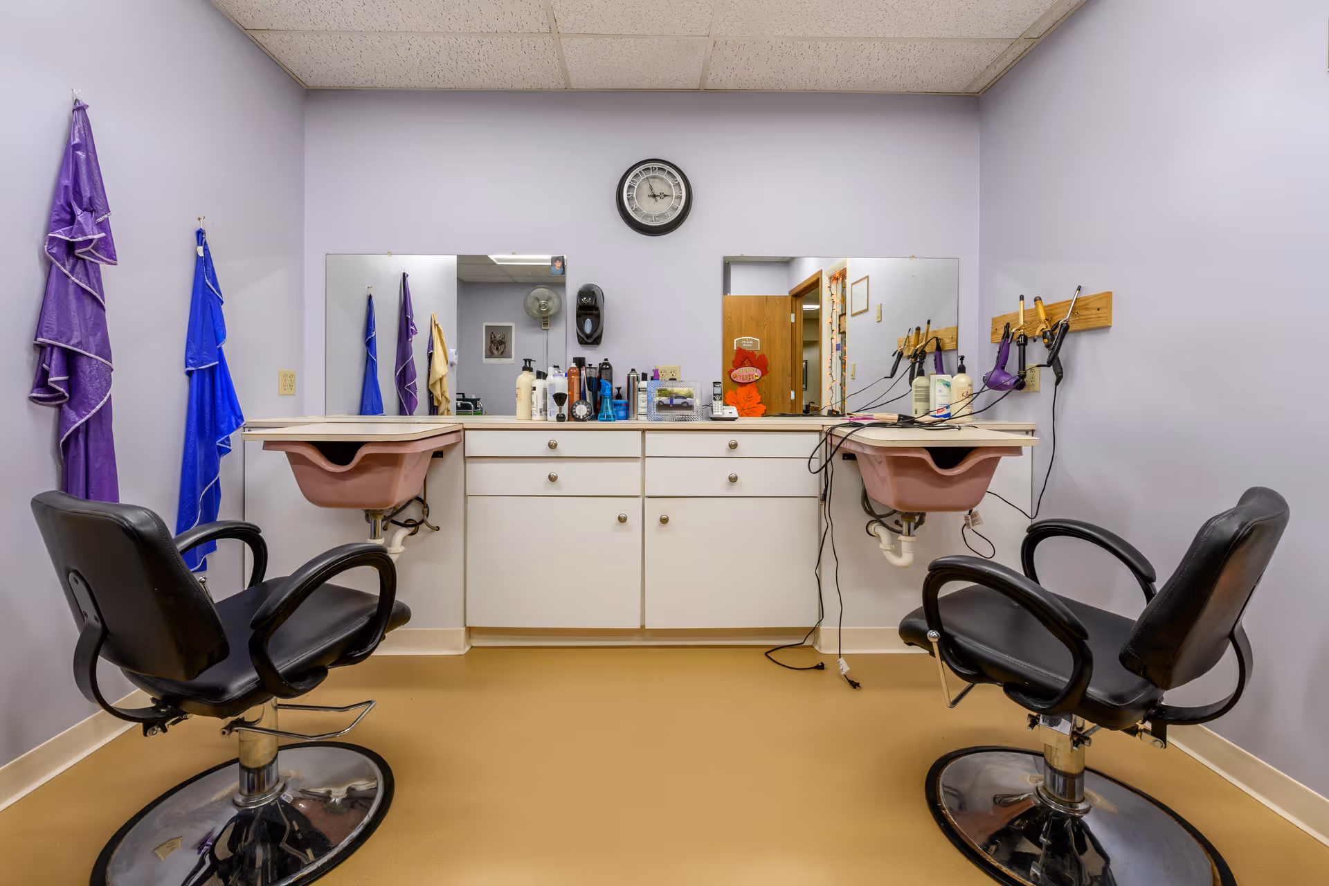 Interior view of a small hair salon area with two black salon chairs facing a counter with two pink sinks. Above the counter are two mirrors and a clock on the wall. Various hair care products and tools are placed on the counter and mounted on the wall. Purple and blue towels hang on hooks on the left wall.