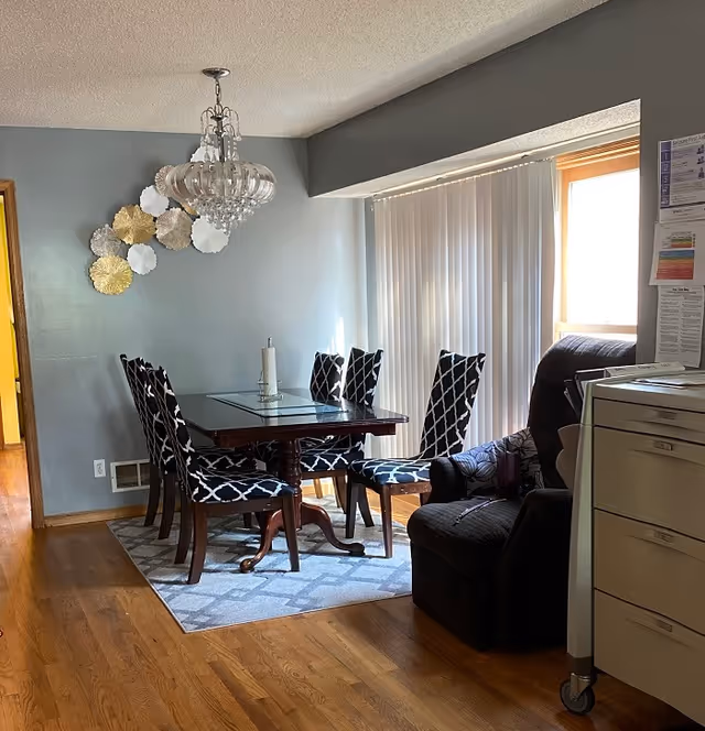 Dining area with a glass-top table surrounded by patterned chairs, a chandelier overhead, a recliner, and vertical blinds over a sliding door.
