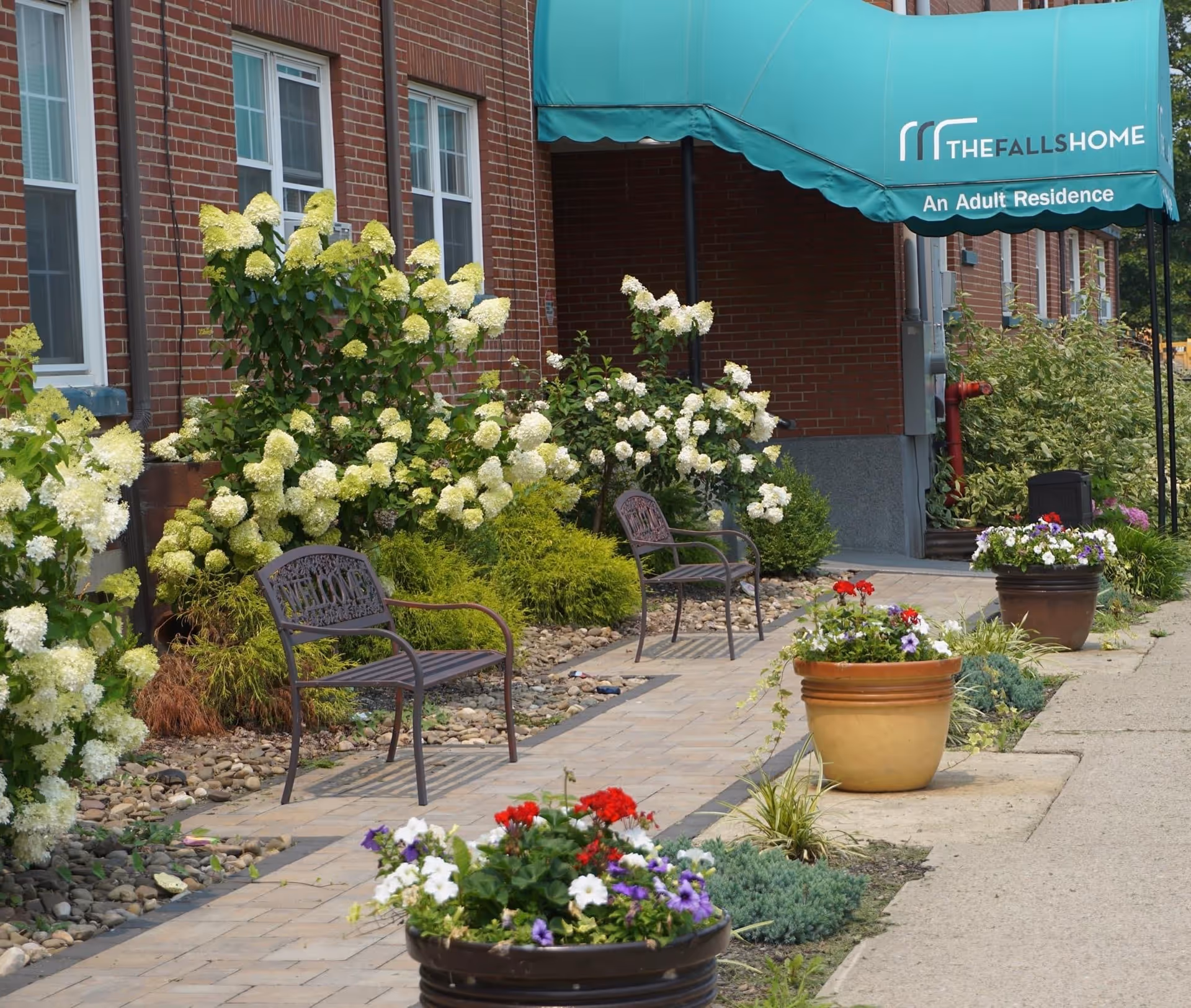 Outdoor seating area with two metal benches along a paved walkway, surrounded by lush green bushes and blooming white and colorful flowers in pots, next to a brick building with windows and a green awning that reads 'THE FALLS HOME An Adult Residence'.