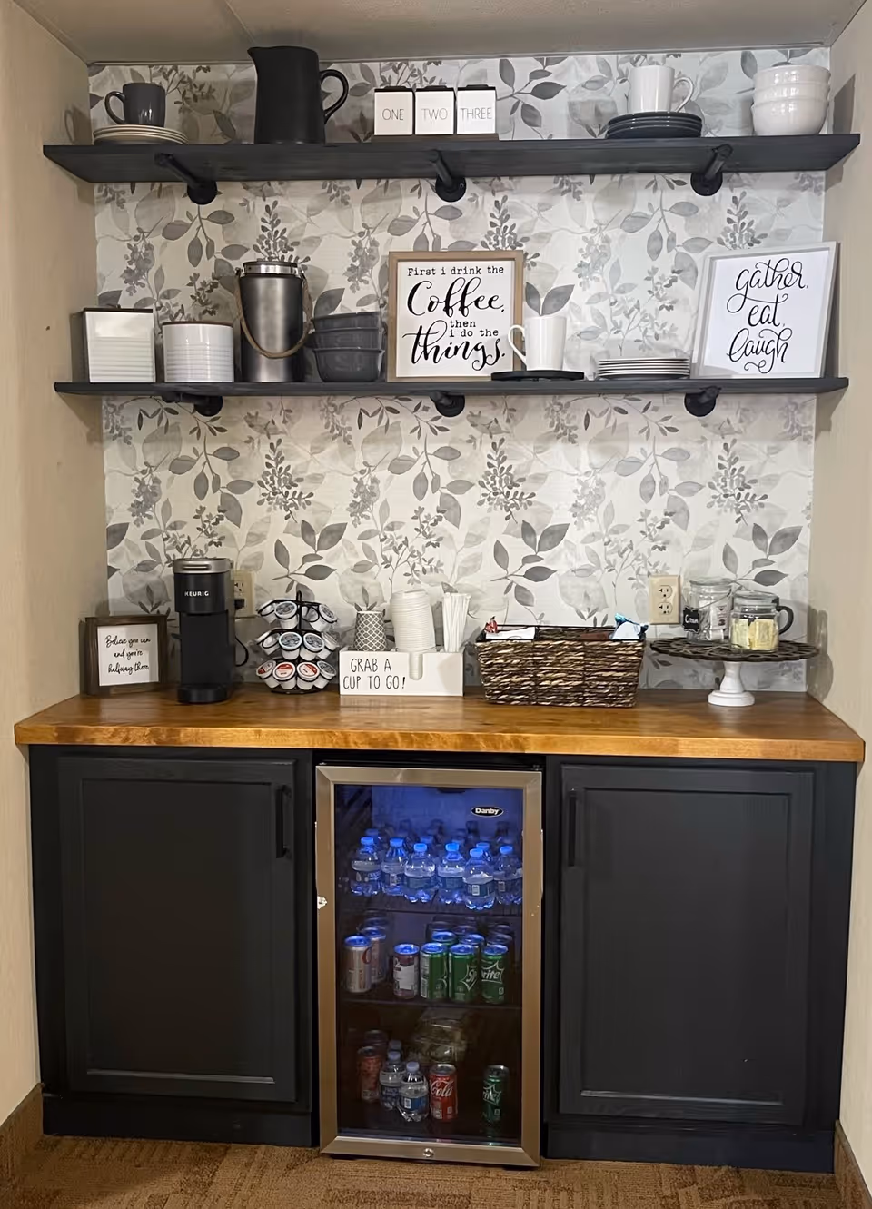 A beverage station with two dark wooden shelves mounted on a floral patterned wall. The shelves hold various cups, bowls, plates, and decorative signs with quotes about coffee and gathering. Below the shelves is a wooden countertop with a Keurig coffee maker, a rotating pod holder, disposable cups, stirrers, a basket with packets, and a small refrigerator stocked with bottled water and soda cans. The cabinetry below the countertop is dark-colored with two closed doors on either side of the refrigerator.