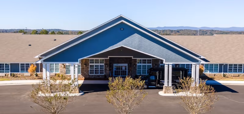 Front exterior of a single-story senior living building with a covered entrance/porte-cochere and a shuttle van parked in front.