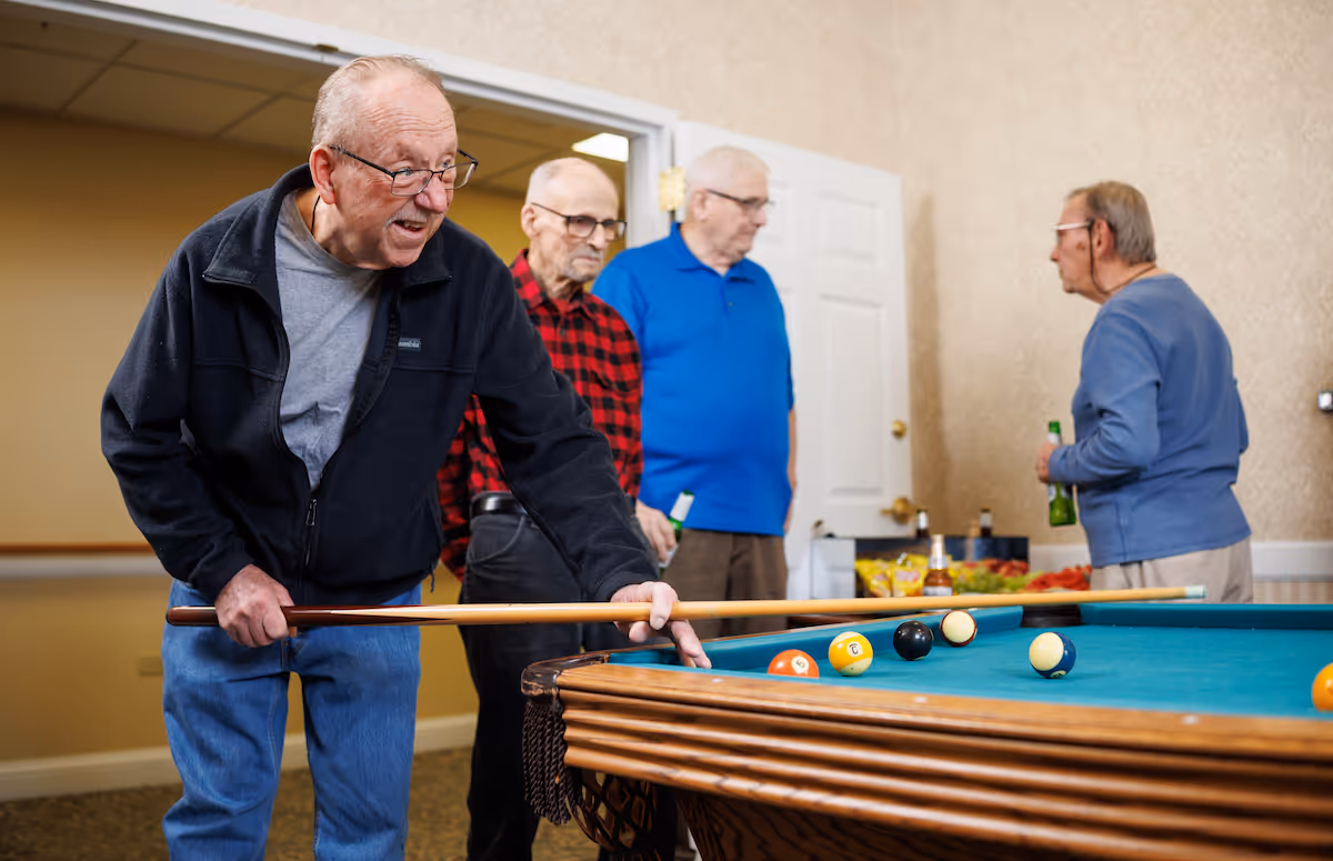 Four elderly men in a room playing pool. One man is leaning over the pool table aiming a shot with a cue stick, while the other three men stand nearby watching and holding drinks. The room has beige walls and a white door in the background.