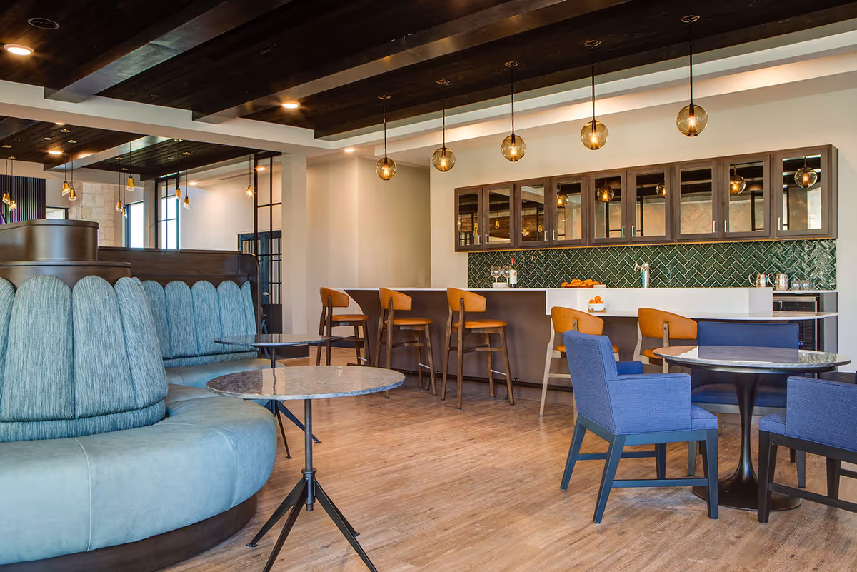 Modern senior living facility lounge area with a curved teal upholstered booth, round marble tables, blue armchairs, and a bar with wooden stools and pendant lighting. The bar has a green tile backsplash and mirrored cabinets above.