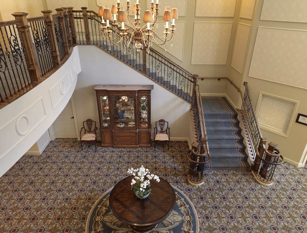 Elevated view of an elegant senior living lobby with a round table and flowers, patterned carpet, a wooden display cabinet, and a grand staircase with wrought-iron railings under a chandelier.