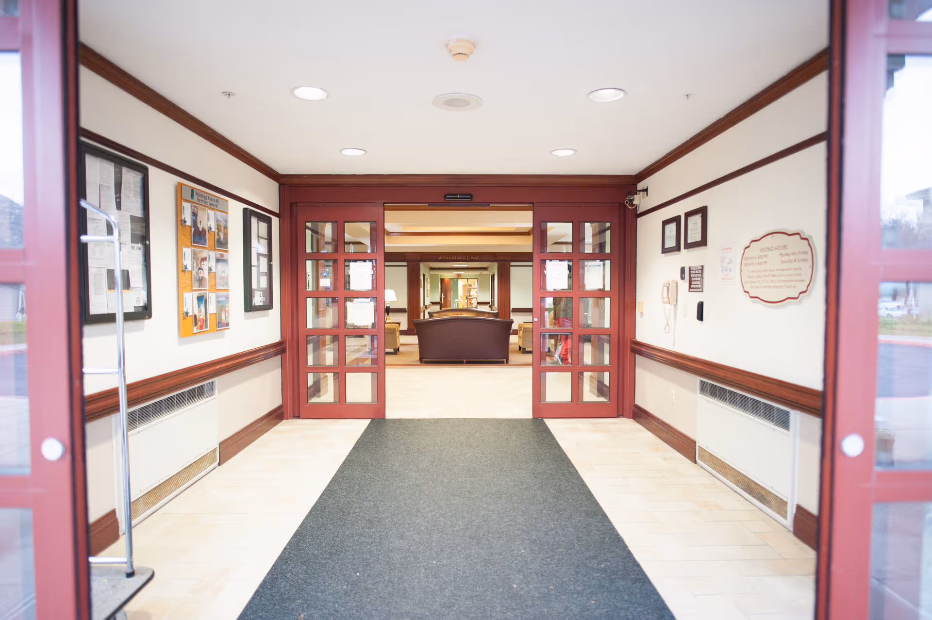 Entrance hallway of EverGreen Commons Rehabilitation and Nursing Center with glass double doors leading to a lounge area furnished with sofas. The hallway has bulletin boards and framed notices on the walls, beige tiled floor with a dark carpet runner, and wood trim accents.