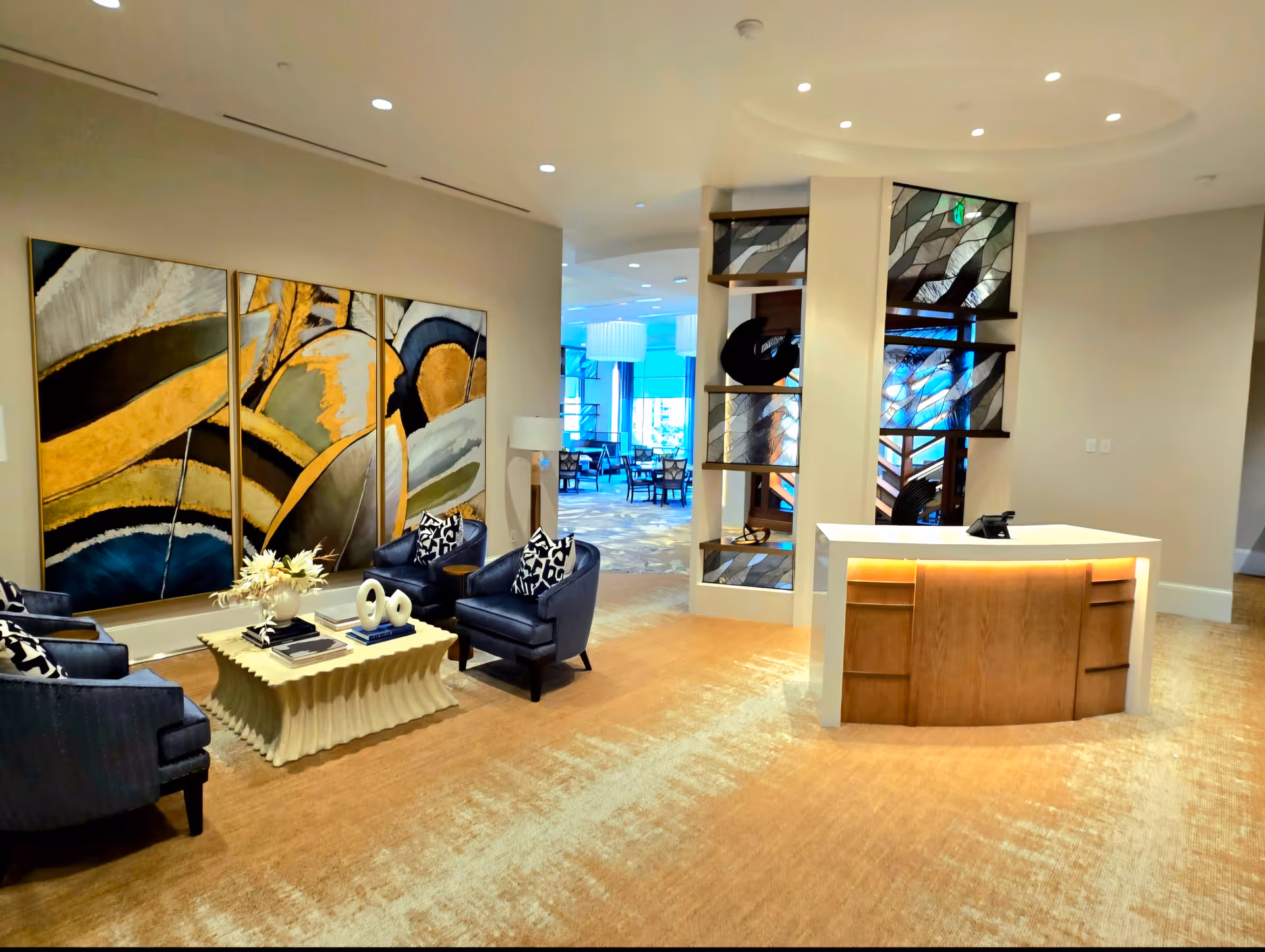 A modern and stylish interior common area with a seating arrangement consisting of four dark blue armchairs around a cream-colored coffee table adorned with decorative items and books. Behind the seating area is a large three-panel abstract painting with gold, black, and gray tones. To the right, there is a reception desk with a wooden front and a white countertop, illuminated from below. Behind the desk is a decorative partition with artistic glass panels and shelves holding abstract sculptures. In the background, a dining area with tables and chairs is visible through an open doorway.