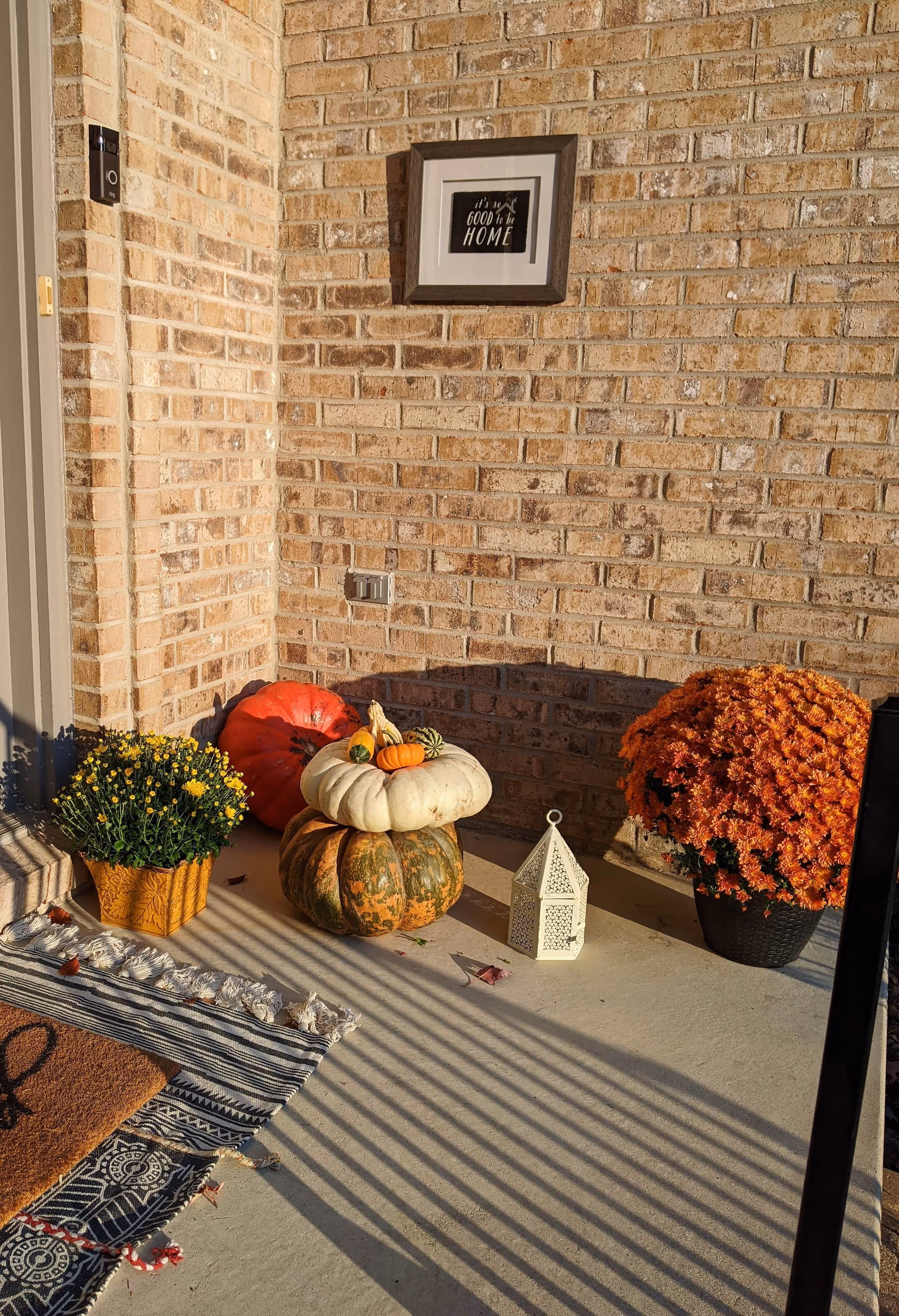 Front porch with a brick wall decorated with stacked pumpkins, potted mums, a lantern, and a welcome mat.