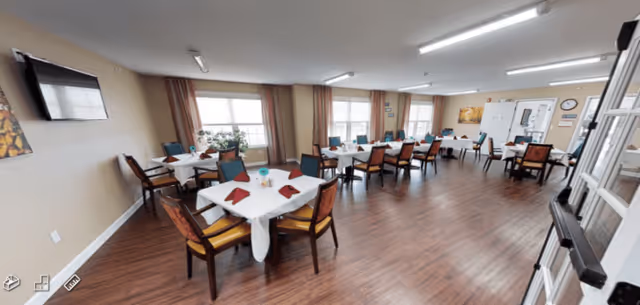 A spacious dining room with multiple tables covered with white tablecloths and set with folded red napkins. Each table is surrounded by wooden chairs with cushioned seats. The room has large windows with curtains allowing natural light to enter, wood flooring, and a wall-mounted television.